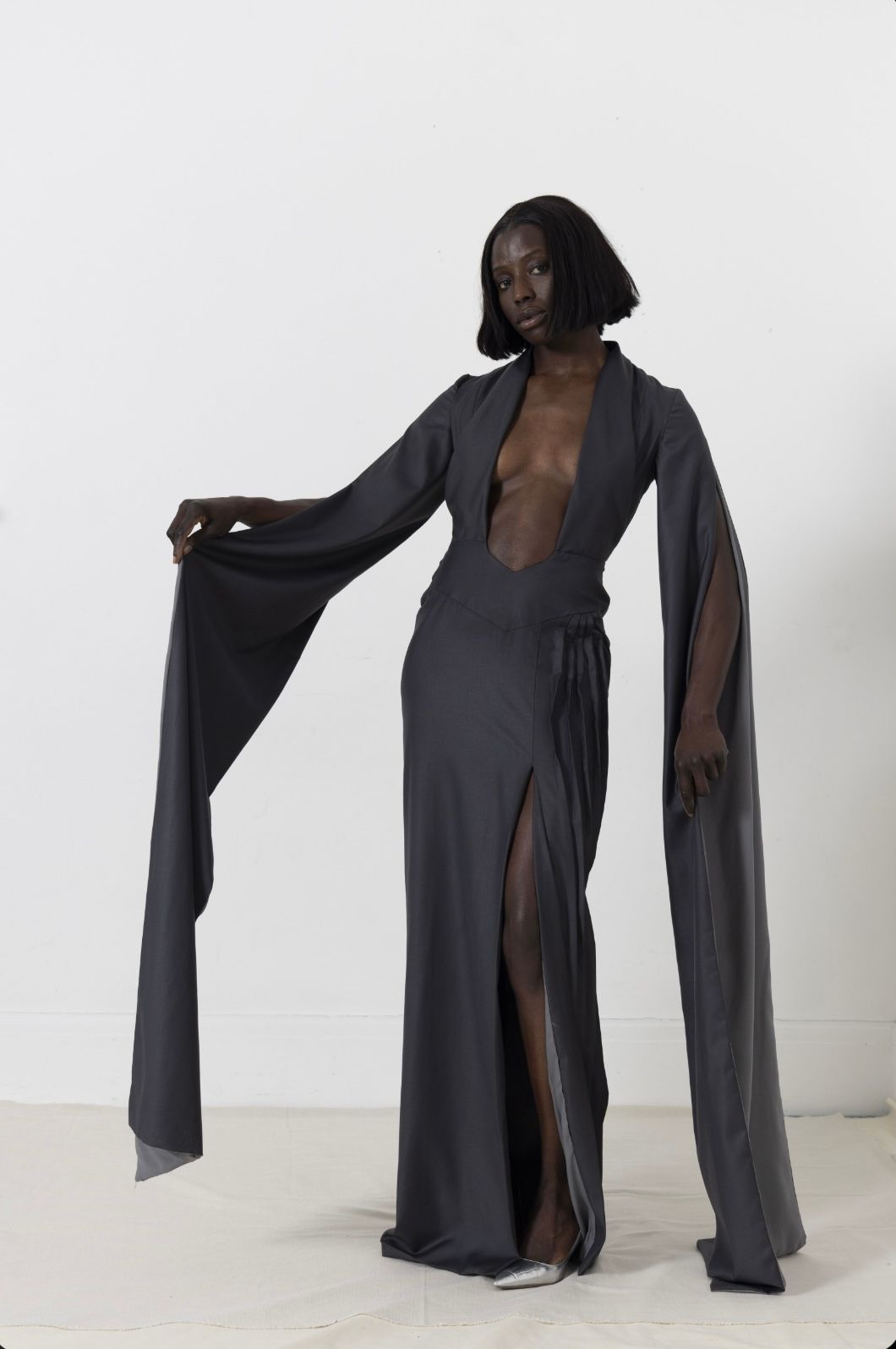 Woman in a dark gray gown with long sleeves and a plunging neckline, stands against a white background.
