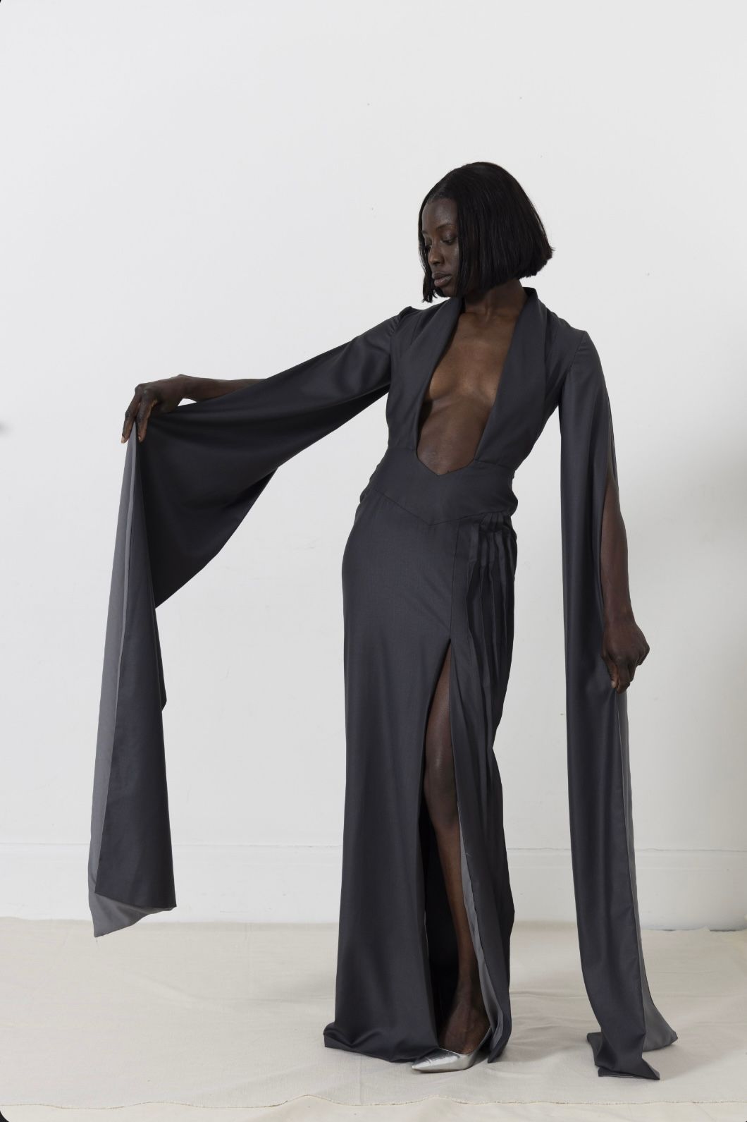 Woman in a dark gray dress with long sleeves and a high slit, posing against a white background.