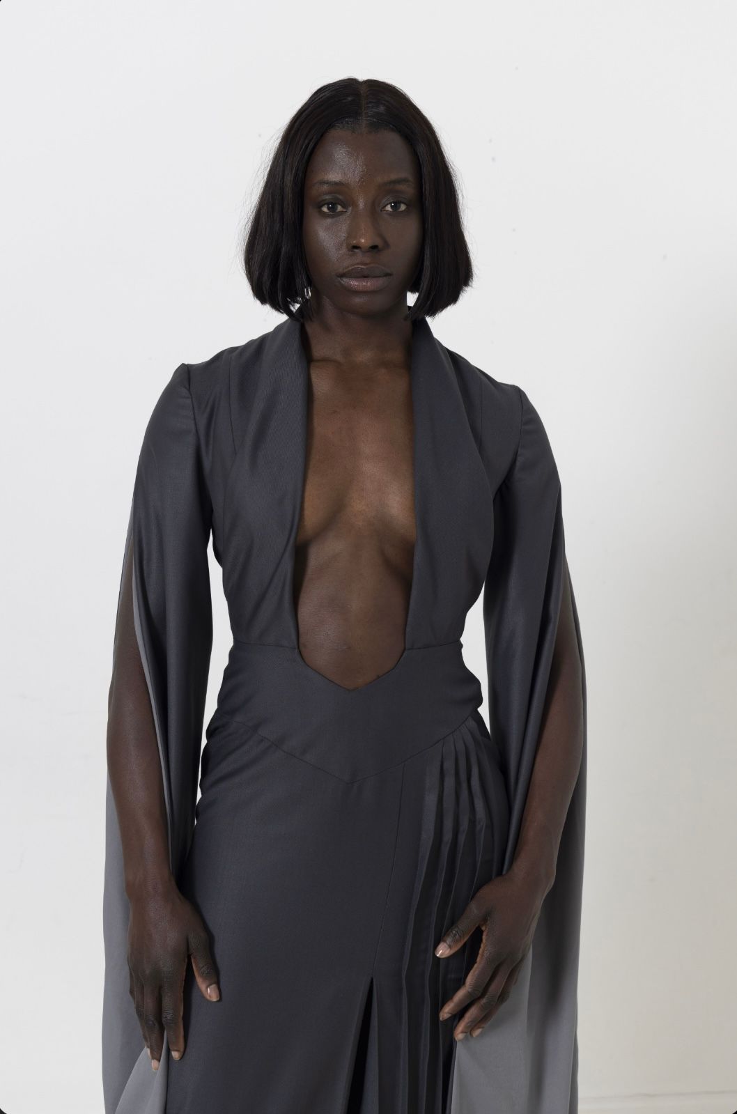 A dark-skinned woman with short dark hair wears a plunging grey dress, standing against a white background.