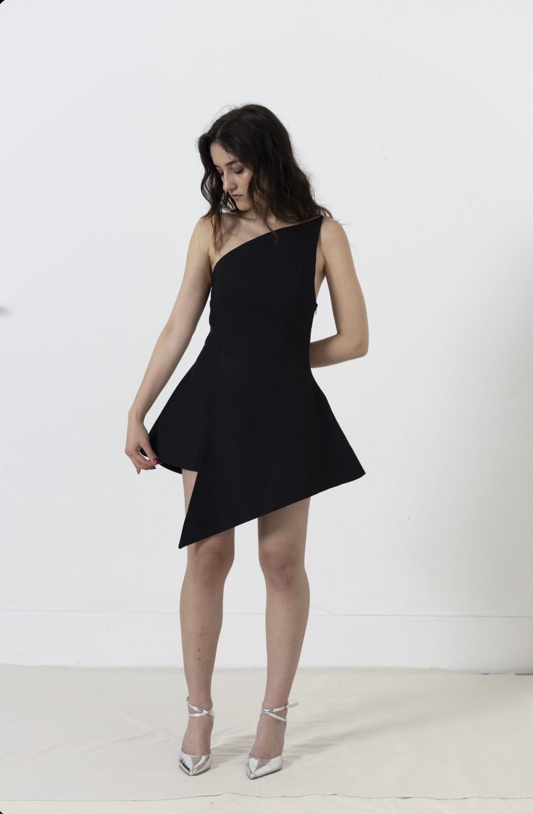 Woman in a black one-shoulder dress and silver heels, posing against a white backdrop.