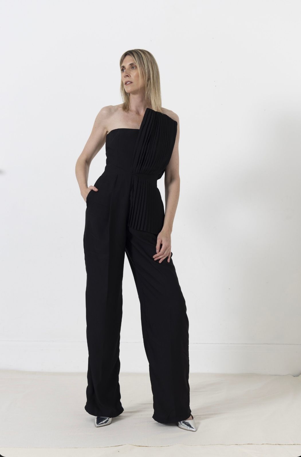 Woman in black strapless jumpsuit, standing with hand in pocket, against white wall.