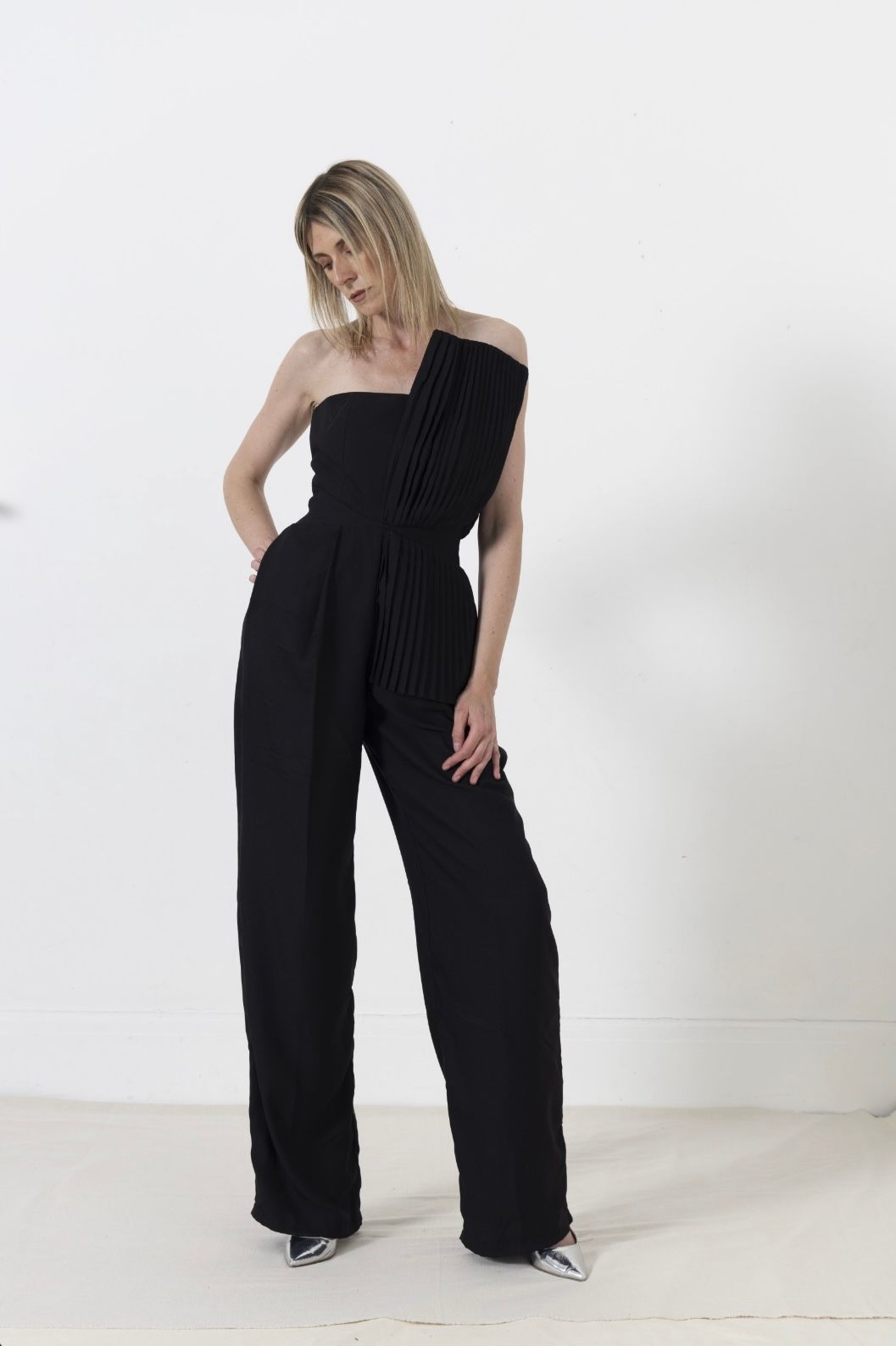 Woman in black strapless jumpsuit, standing with hand in pocket, against white wall.
