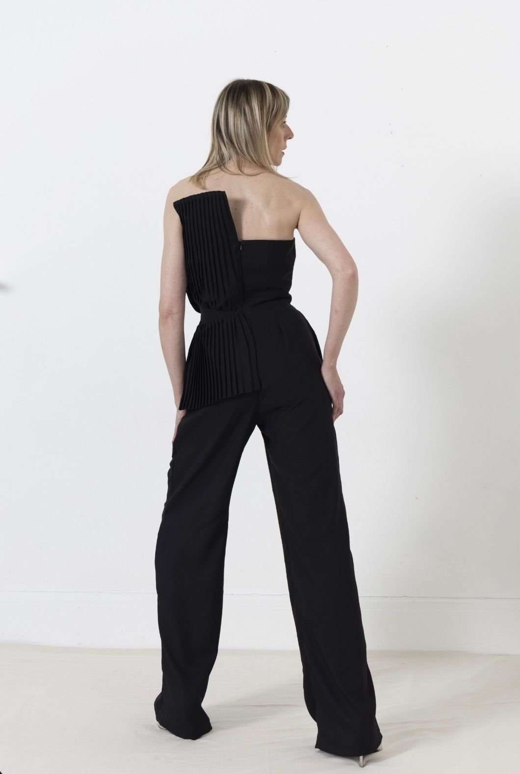 Woman in black jumpsuit, standing against white background, looking over shoulder.