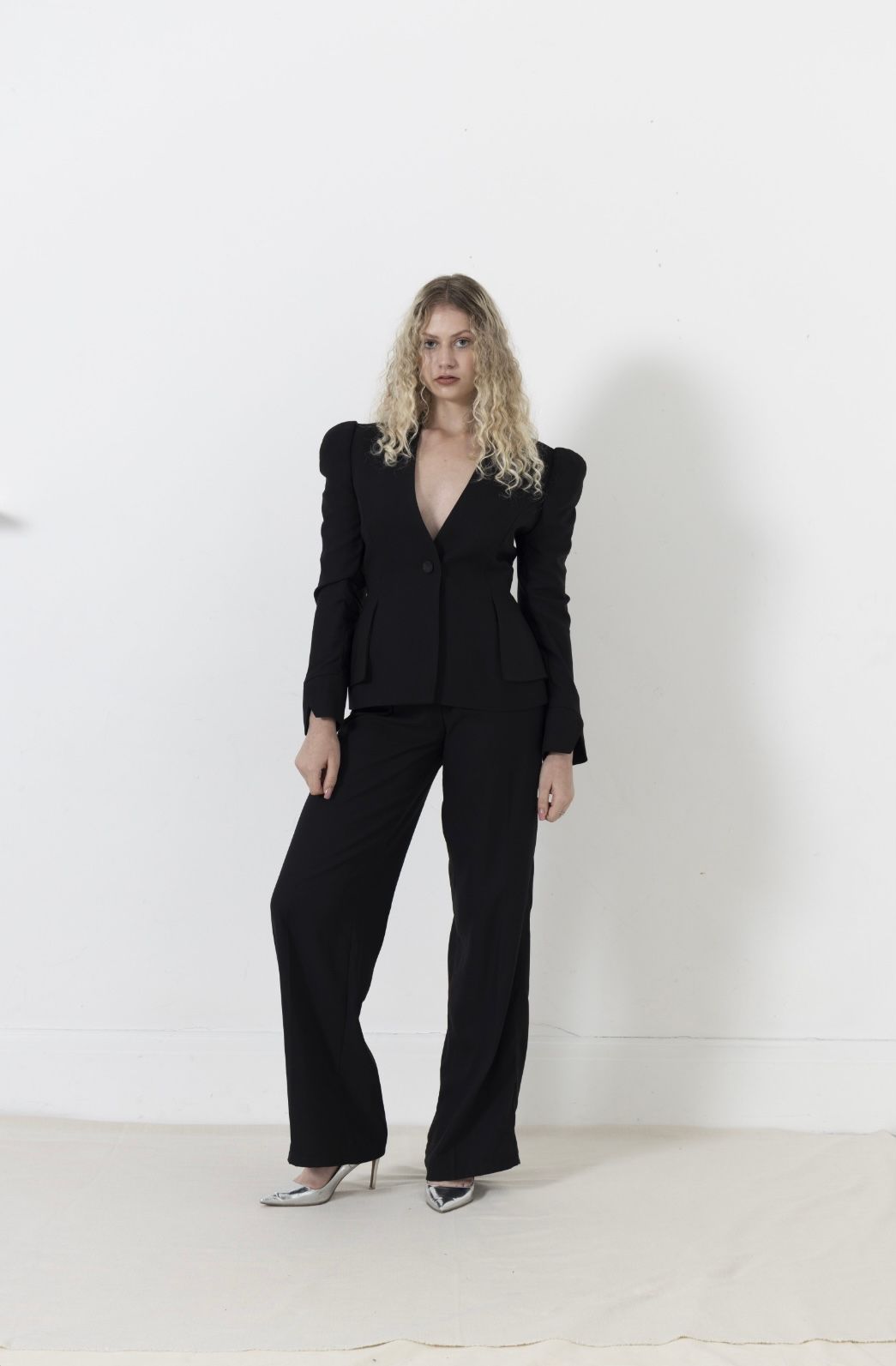 Woman in black suit with structured shoulders, standing against a white wall.