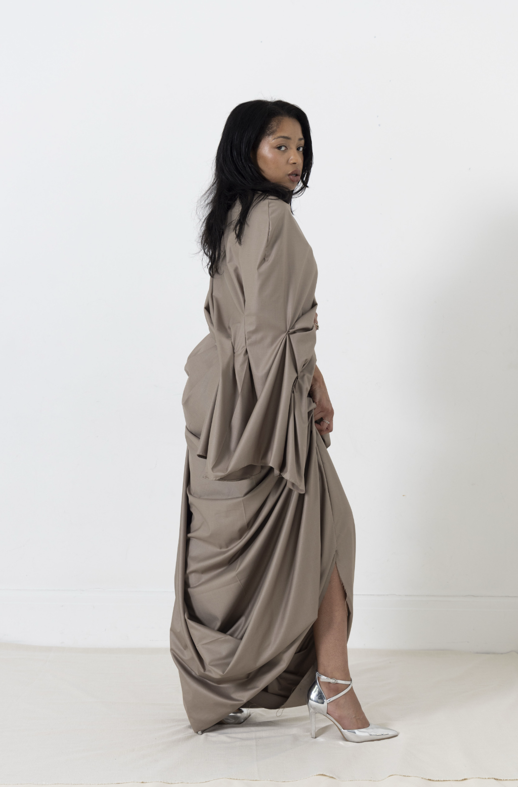 Woman in draped taupe fabric holding up the skirt, standing in a white room, wearing silver heels.