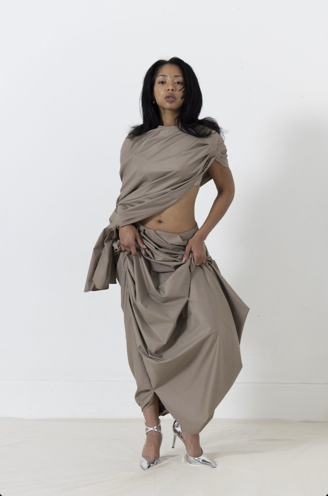 Woman in draped taupe fabric holding up the skirt, standing in a white room, wearing silver heels.