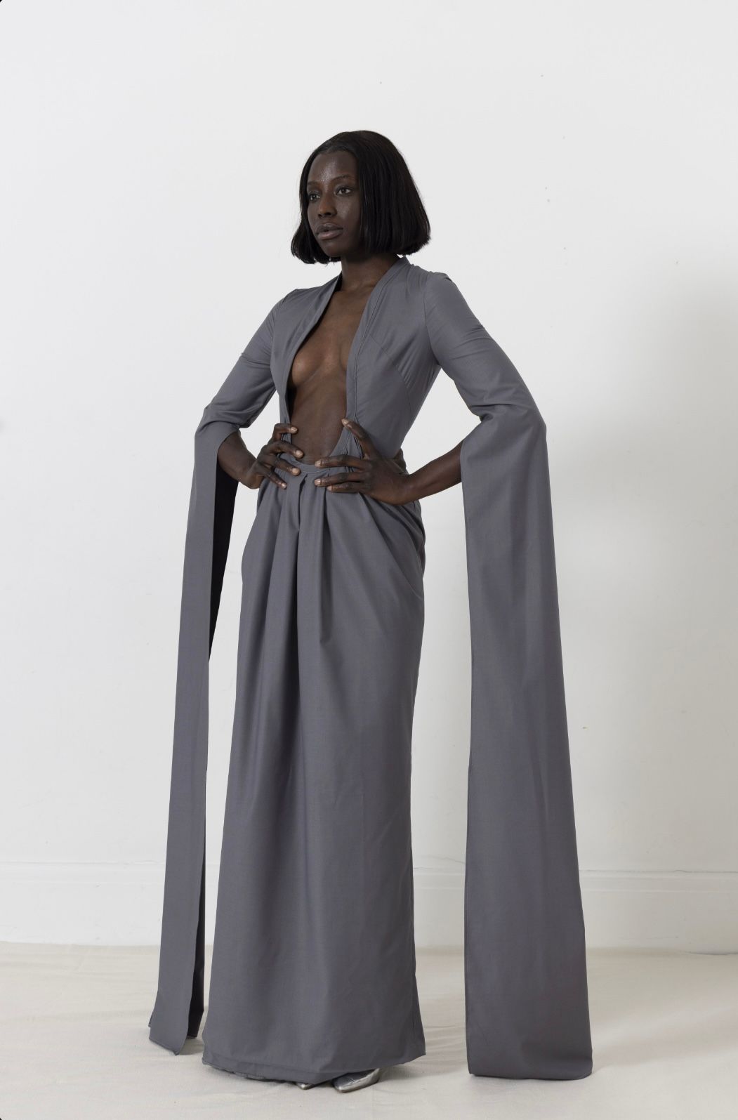 Woman in a gray dress with long, draping sleeves poses against a white background.