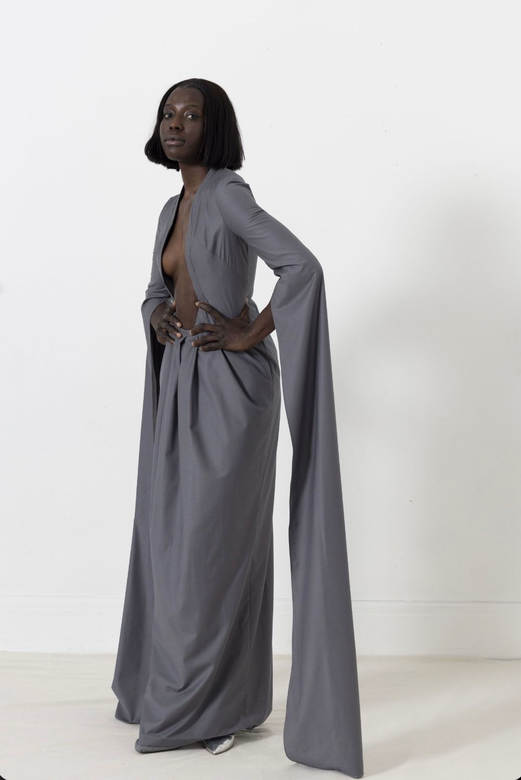 Woman in gray outfit: long skirt, open top, flowing sleeves, hands on hips, against a white wall.
