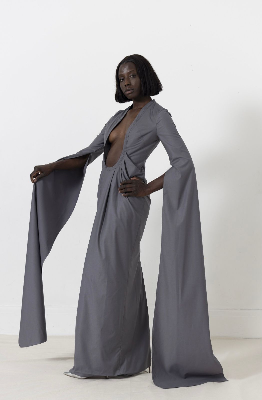 Woman in a long gray dress with plunging neckline and draped sleeves, standing against a white wall.