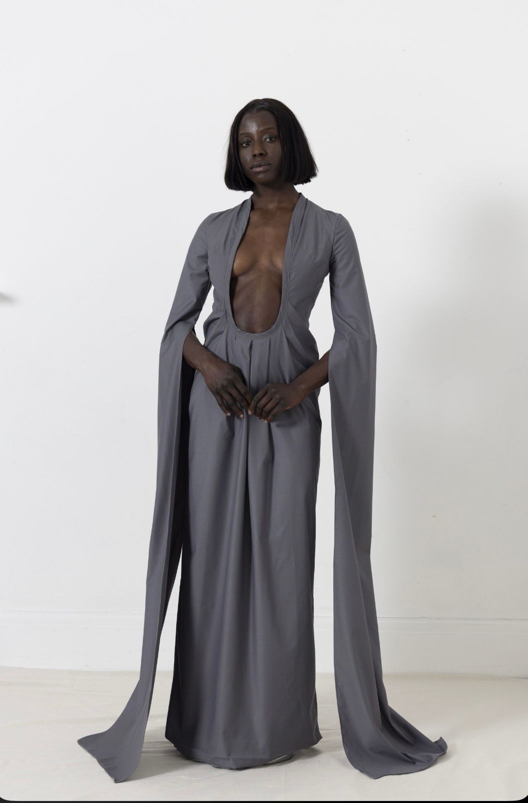 Woman in a long gray dress with plunging neckline and draped sleeves, standing against a white wall.