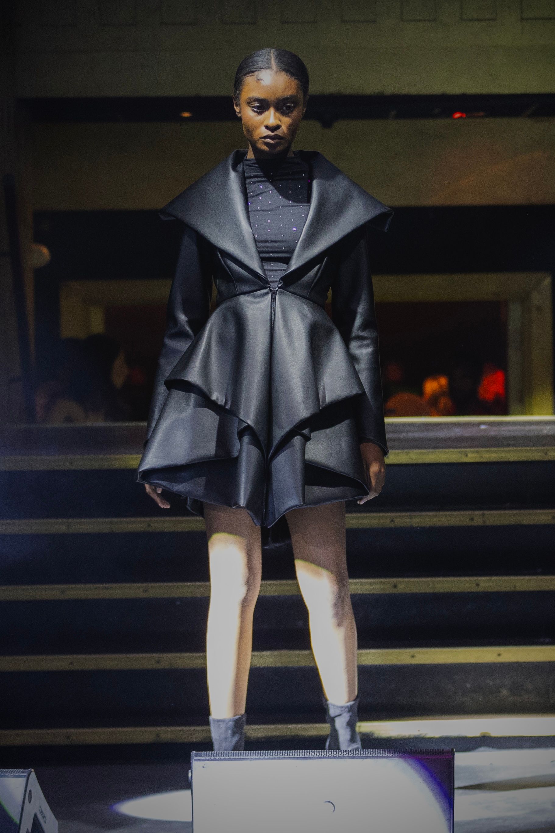Model wearing a black leather jacket with tiered skirt stands on platform in a dimly lit room.