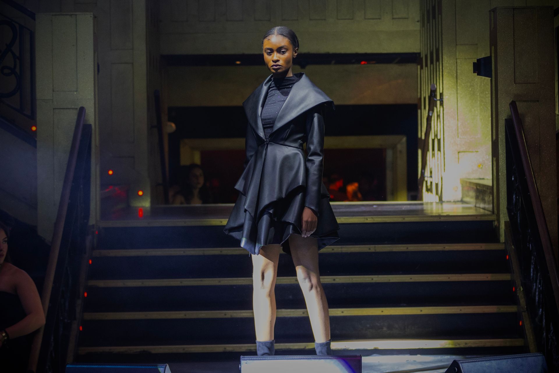 Model in black avant-garde dress walks a runway. Staircase setting.