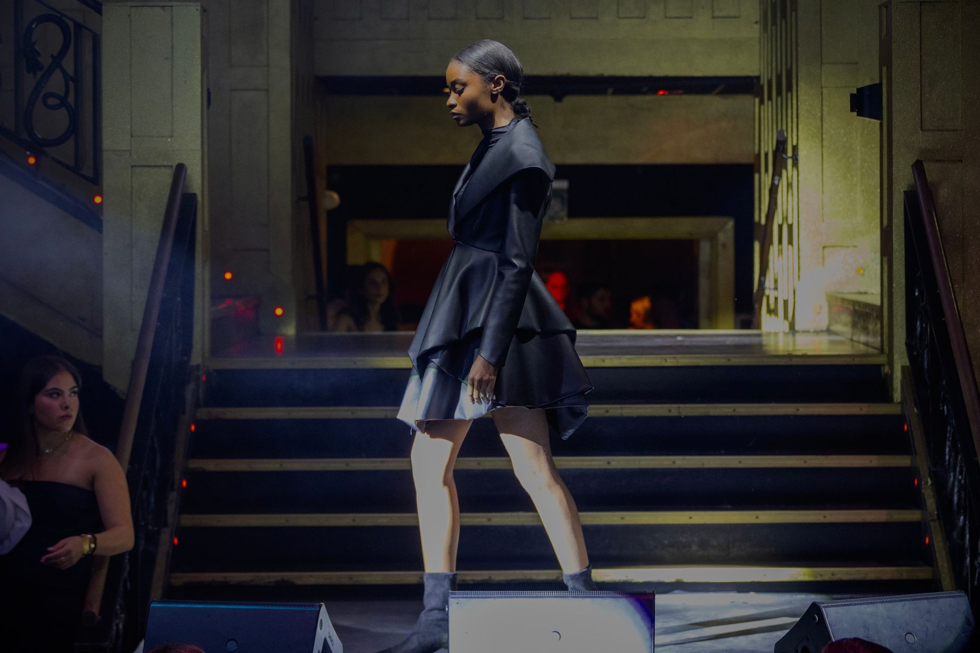 Model in black avant-garde dress walks a runway. Staircase setting.