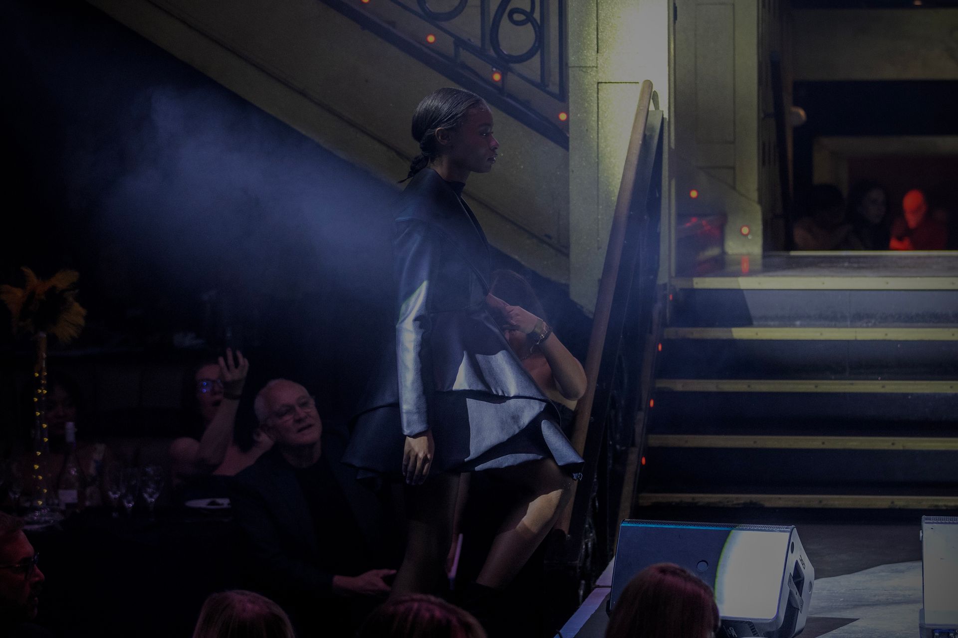 Woman in dark dress descends stairs at an event, spotlighting her with blurred background of people and decor.