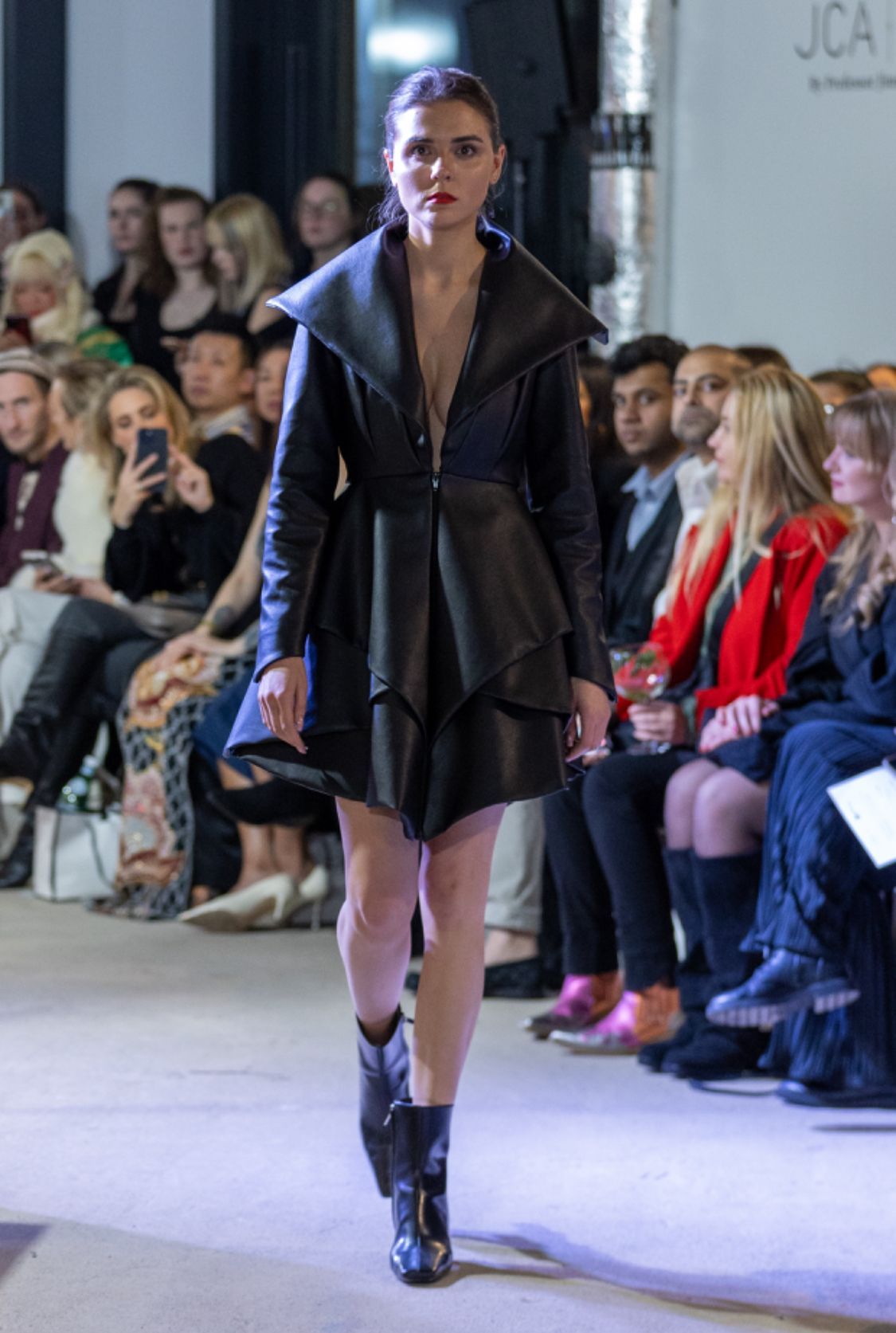 Model in black leather jacket and boots, runway setting.