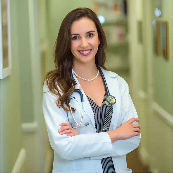 Woman in white coat smiles, arms crossed, in a bright hallway.