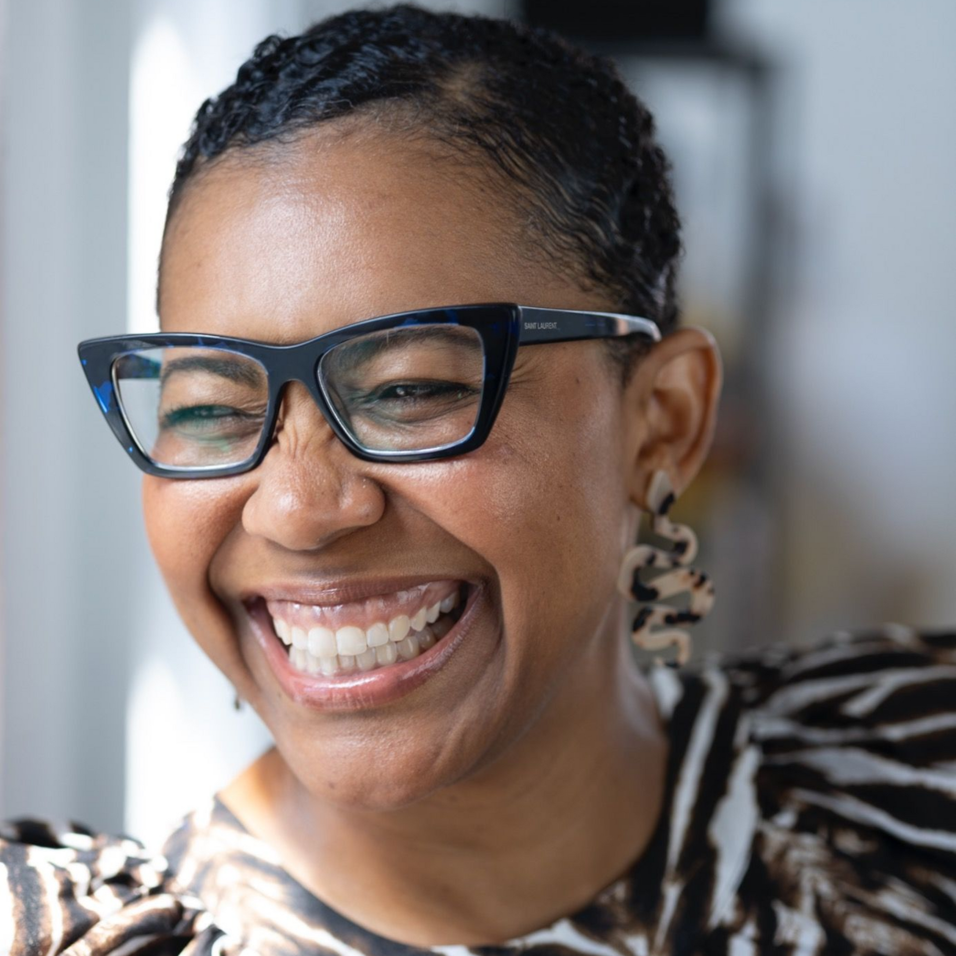 Woman smiling widely, wearing glasses and patterned top, indoors.