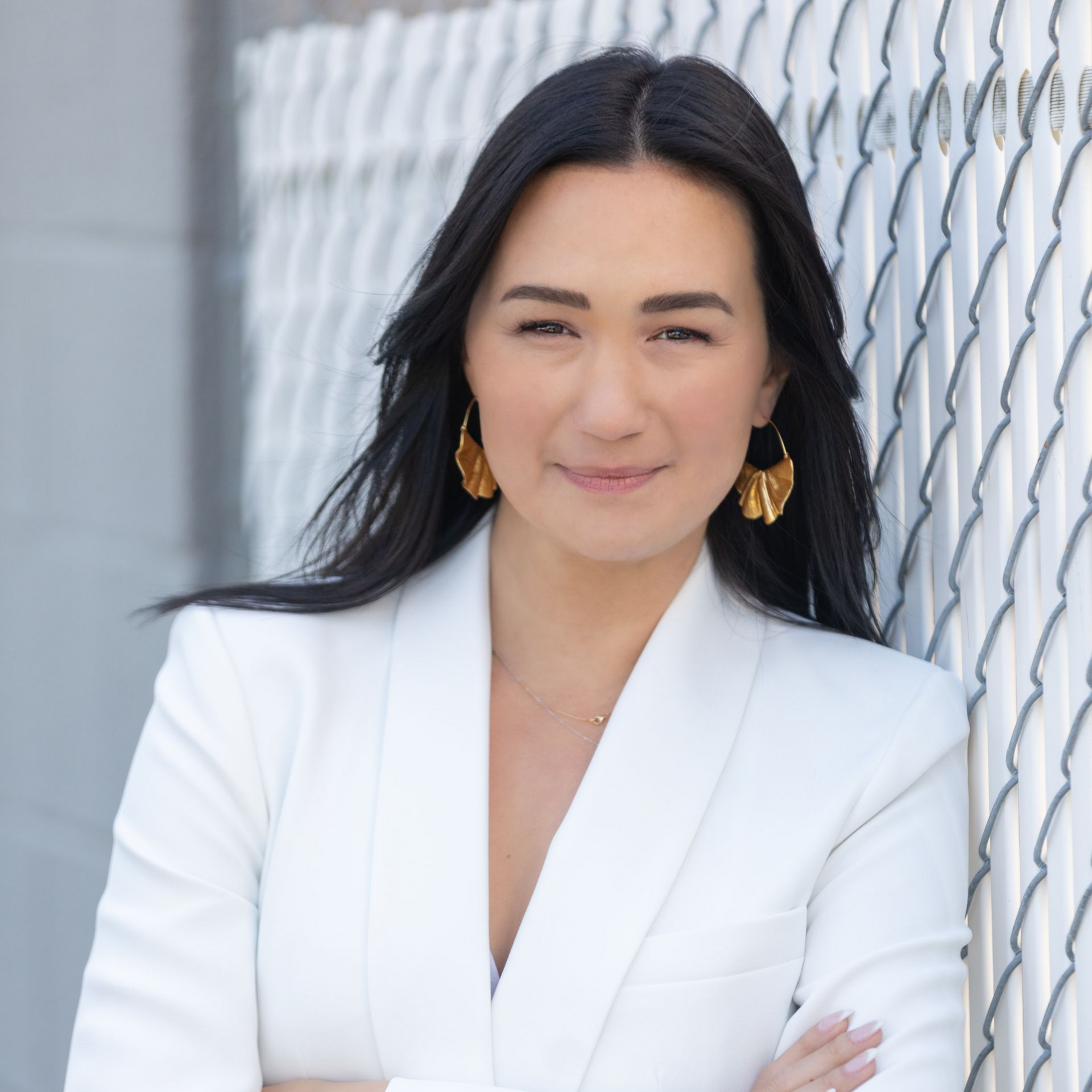 Woman in white blazer, arms crossed, smiling, leaning against a textured metal wall.
