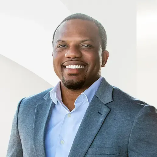 Man in gray blazer smiles; outdoors, bright background.