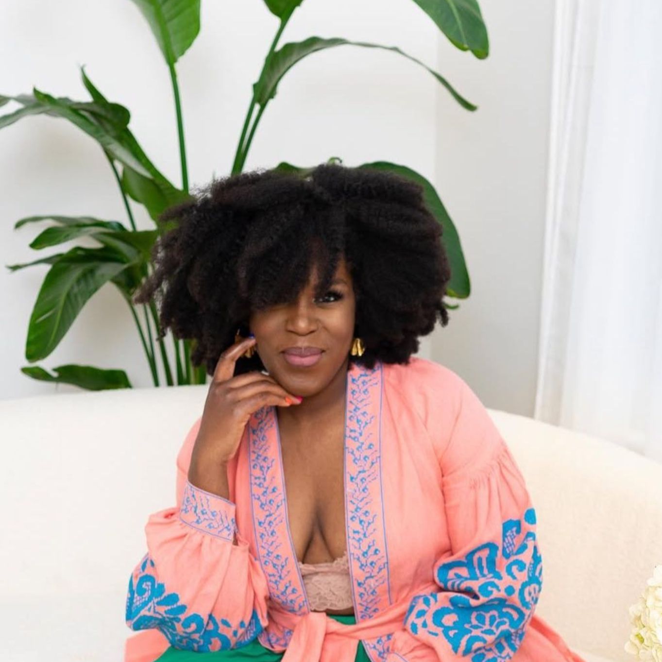 Woman in coral robe, resting chin on hand, looking at camera; indoor setting, foliage in background.