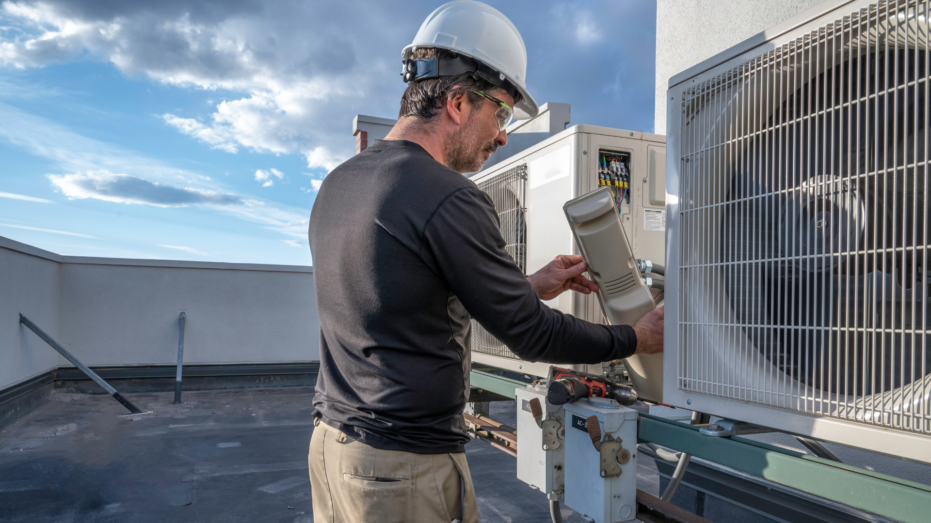 A man wearing a hard hat is working on an air conditioner on the roof of a building.