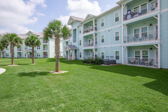 Blue apartment buildings with balconies and palm trees on a grassy lawn under a bright sky.