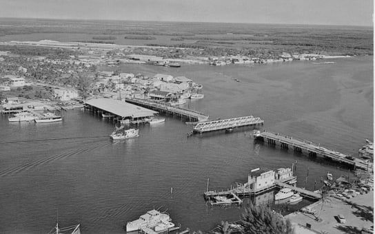 A black and white photo of a harbor with boats and a bridge.