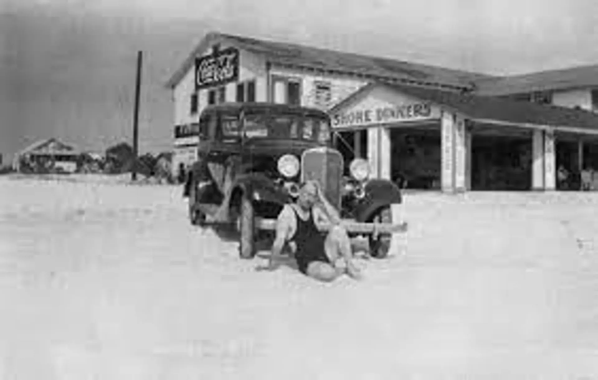 A man in a bathing suit is kneeling next to a car in front of a building.