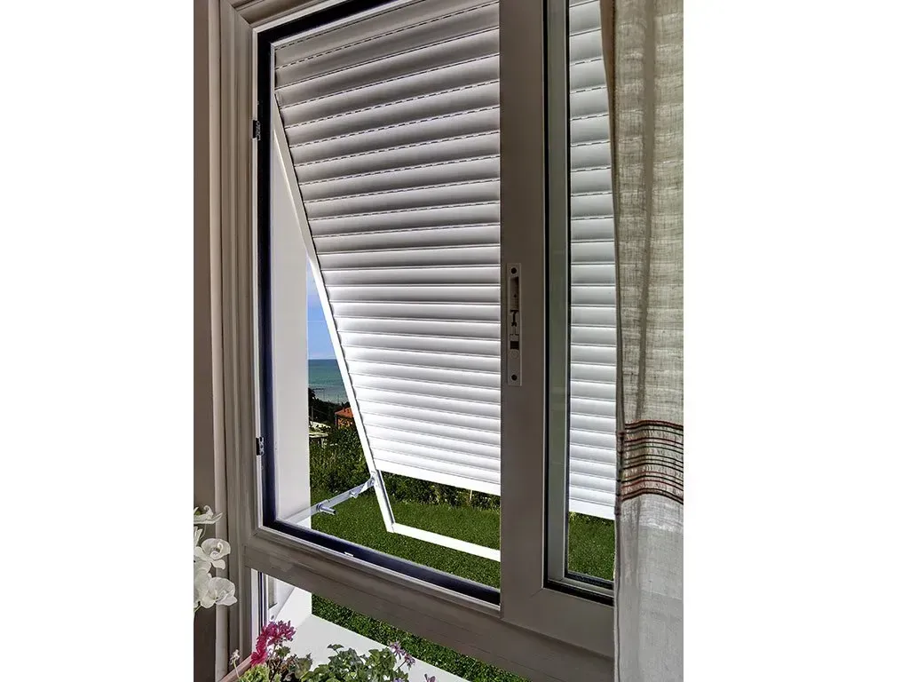 Finestra con persiana avvolgibile bianca parzialmente aperta, che rivela la vista del fogliame verde e dell'acqua.