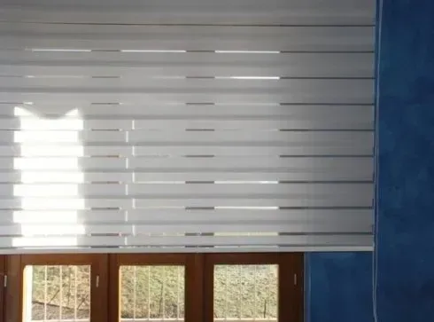 Le tende zebrate bianche coprono parzialmente una finestra con telaio in legno, lasciando filtrare la luce del sole all'interno.