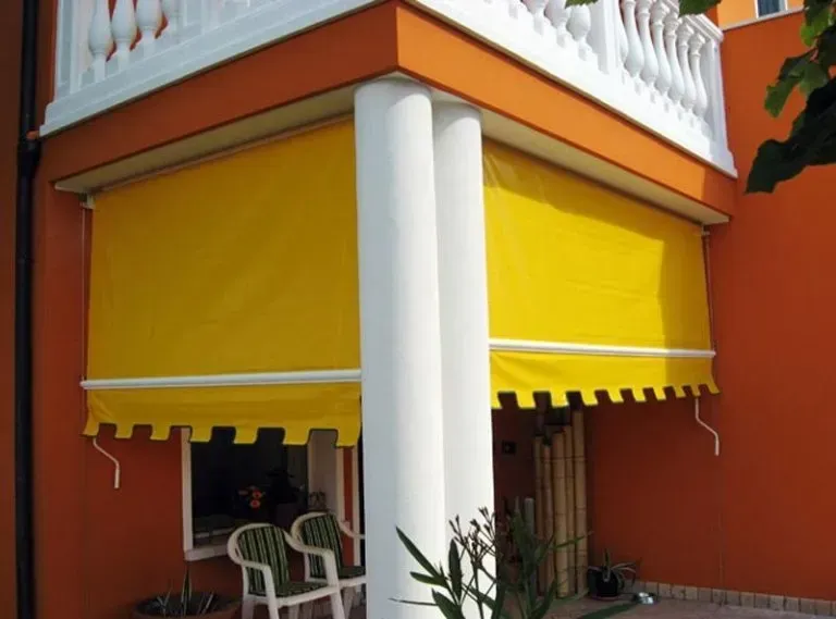 Una tenda da sole gialla ombreggia un angolo di un edificio con colonne bianche e pareti arancioni.