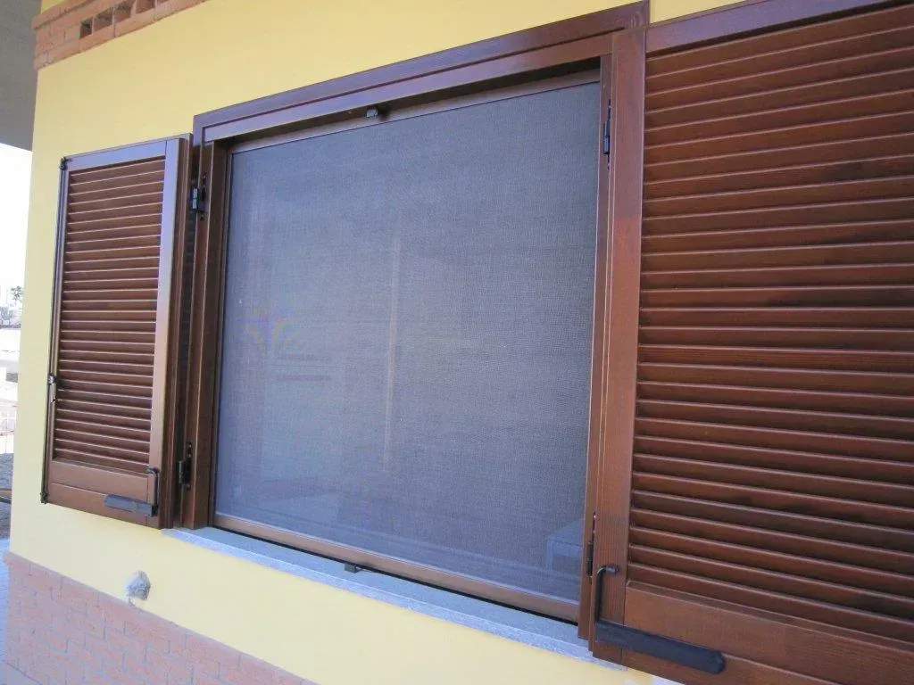 Finestra con persiane marroni e una zanzariera scura su una parete gialla.