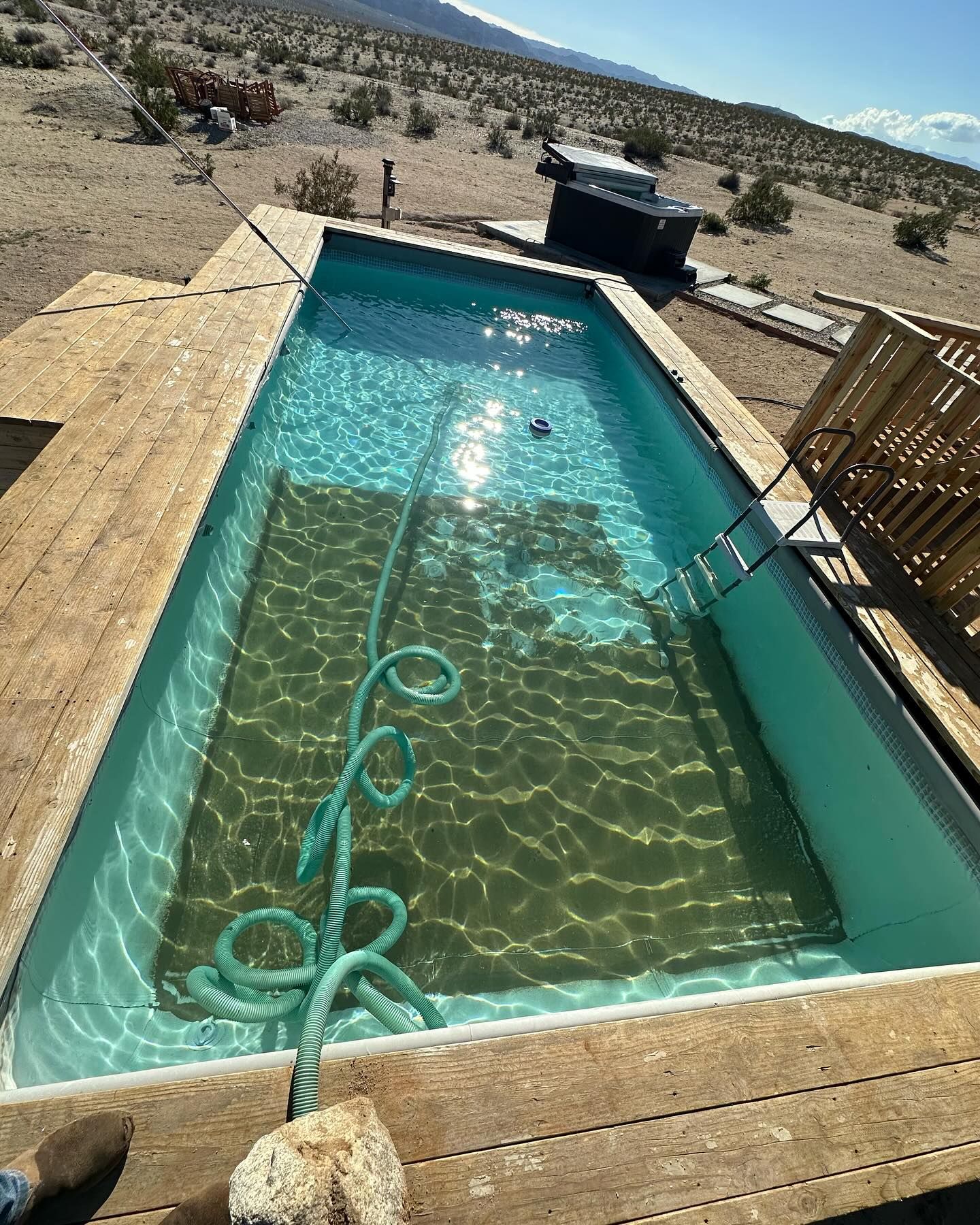 Rectangular pool with wooden deck in a desert setting; a blue hose lies in the water.