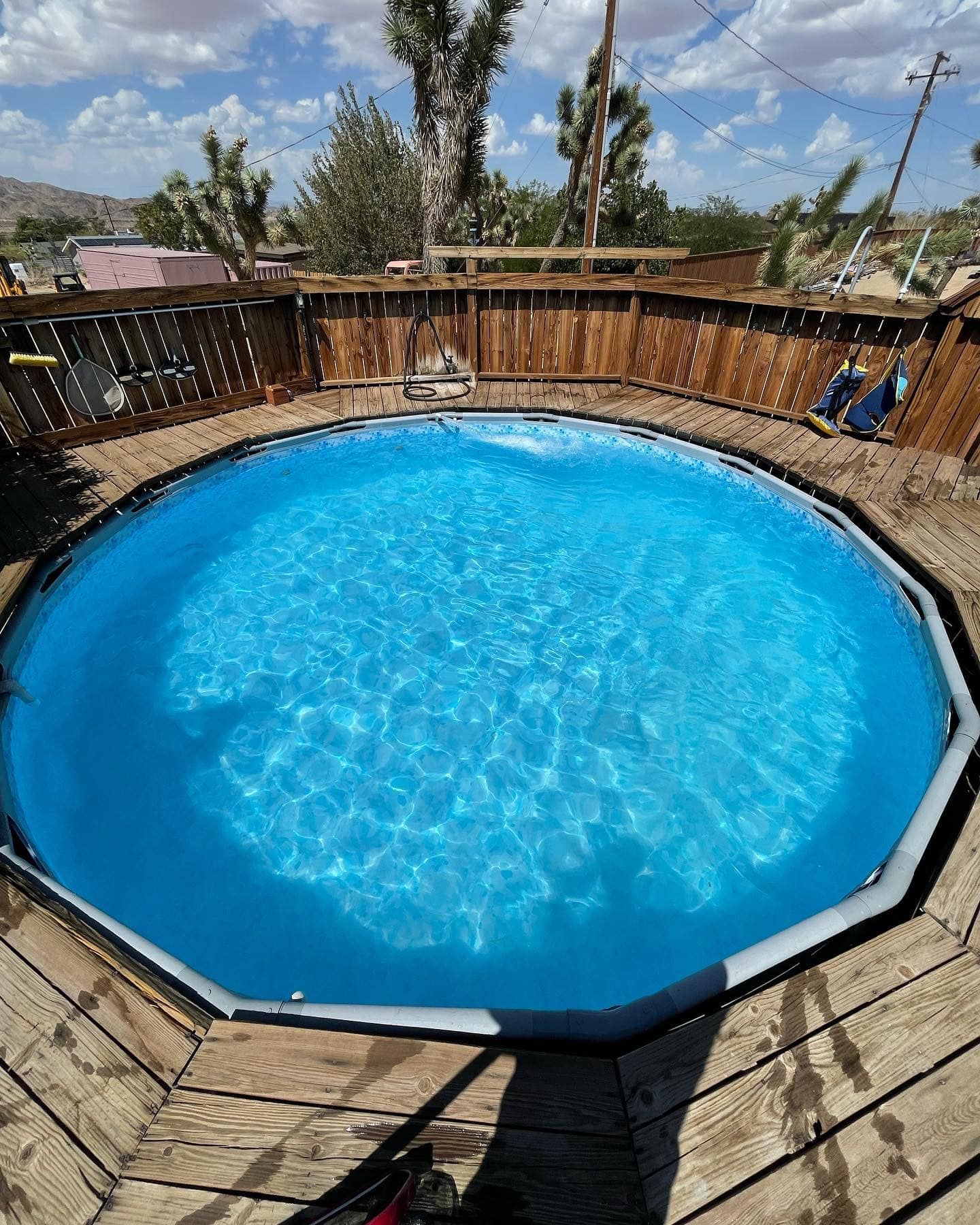 Round blue above-ground swimming pool surrounded by a wooden deck and fence under a sunny sky.