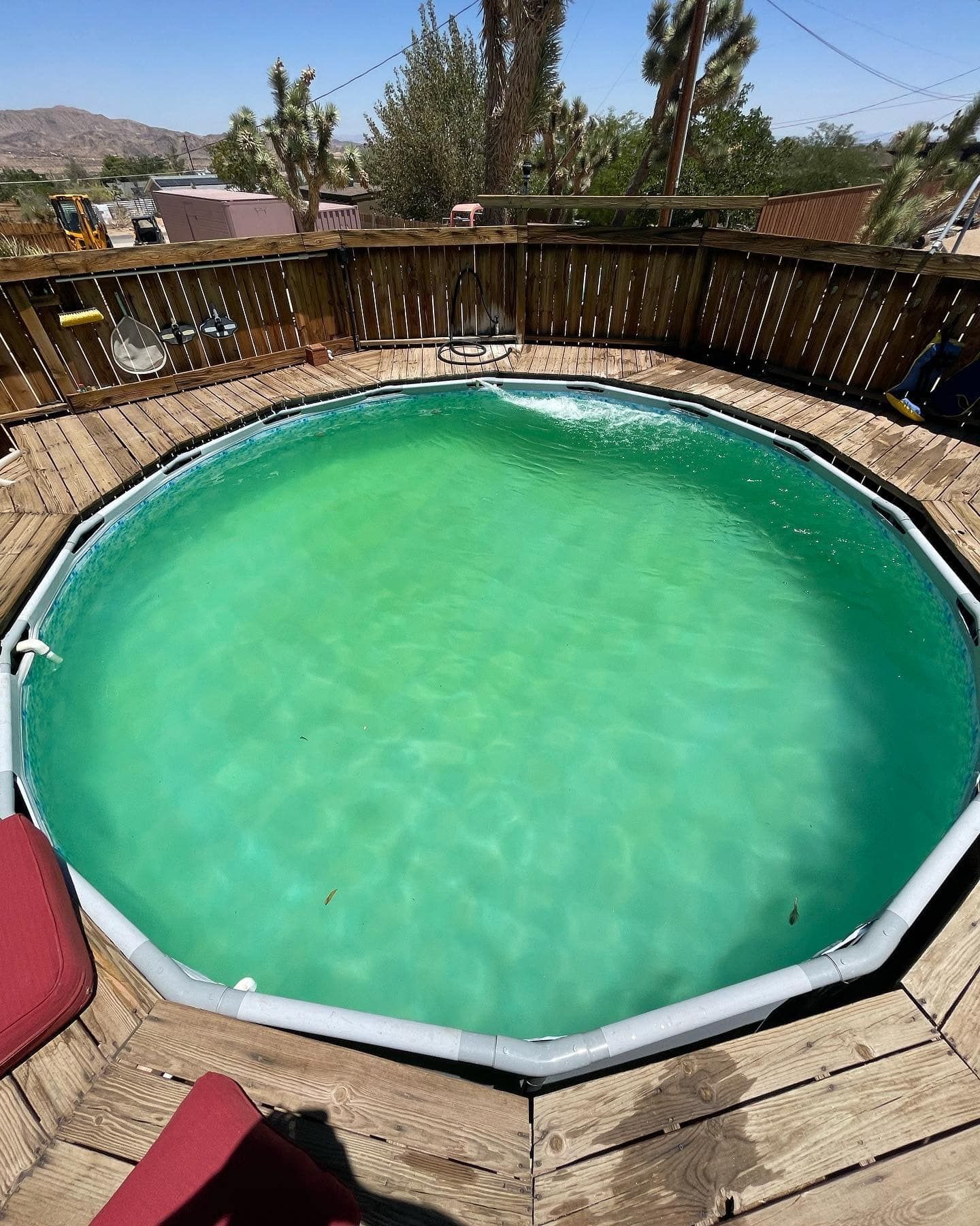 Green above-ground pool surrounded by a wooden deck, under a bright, sunny sky.