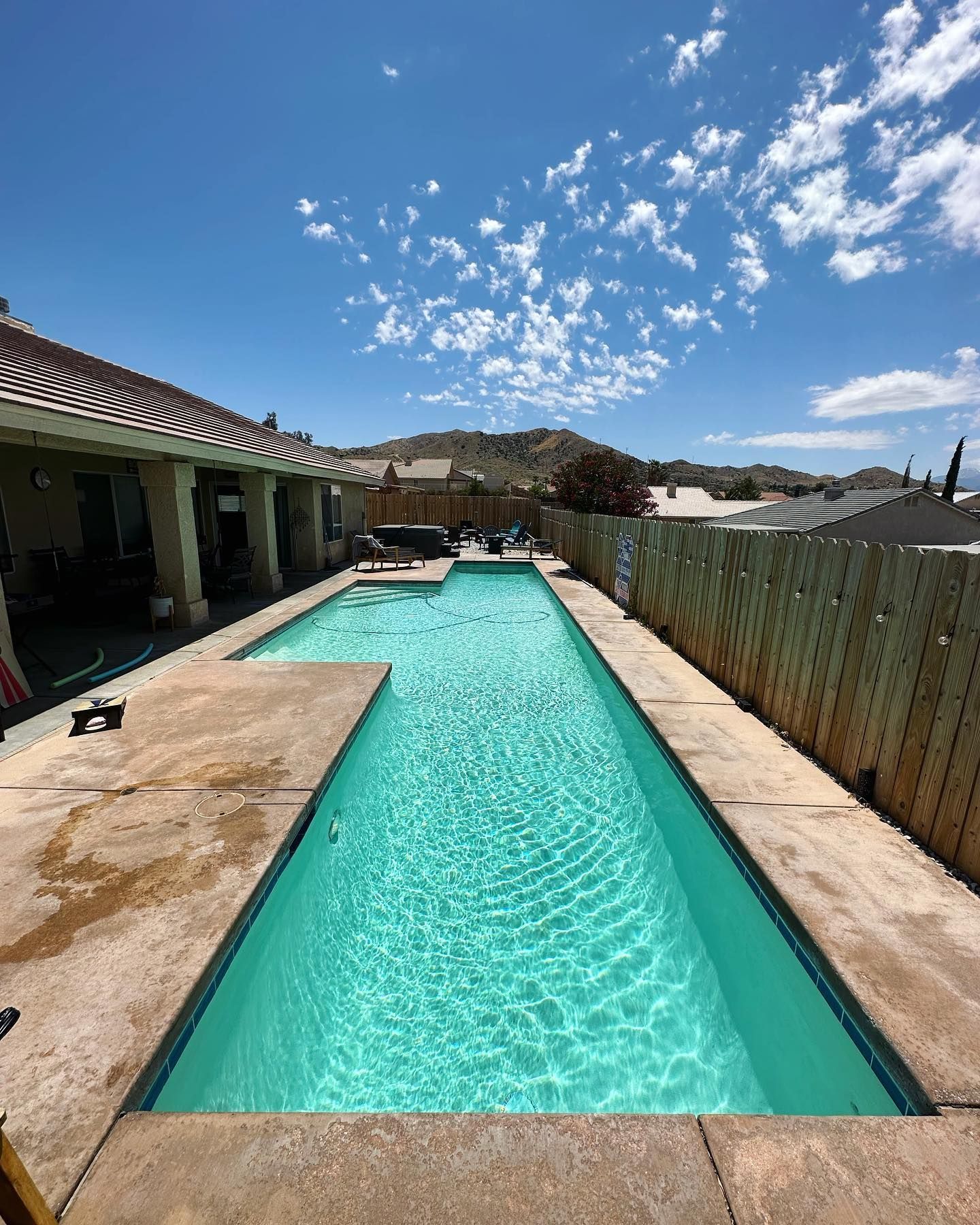 Rectangular swimming pool on a sunny day, next to a house and wooden fence, with mountain in the background.