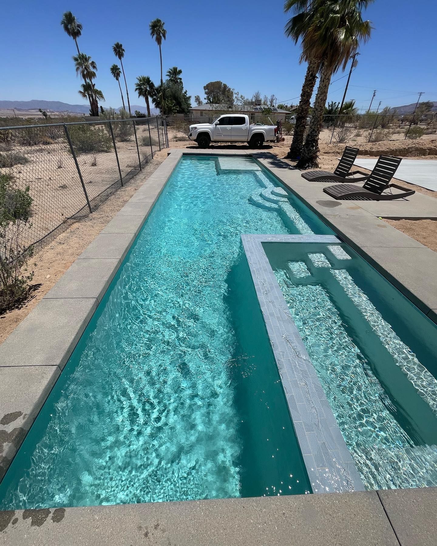 Rectangular turquoise pool with concrete surround. Palm trees and a white truck in the background. Sunny day.
