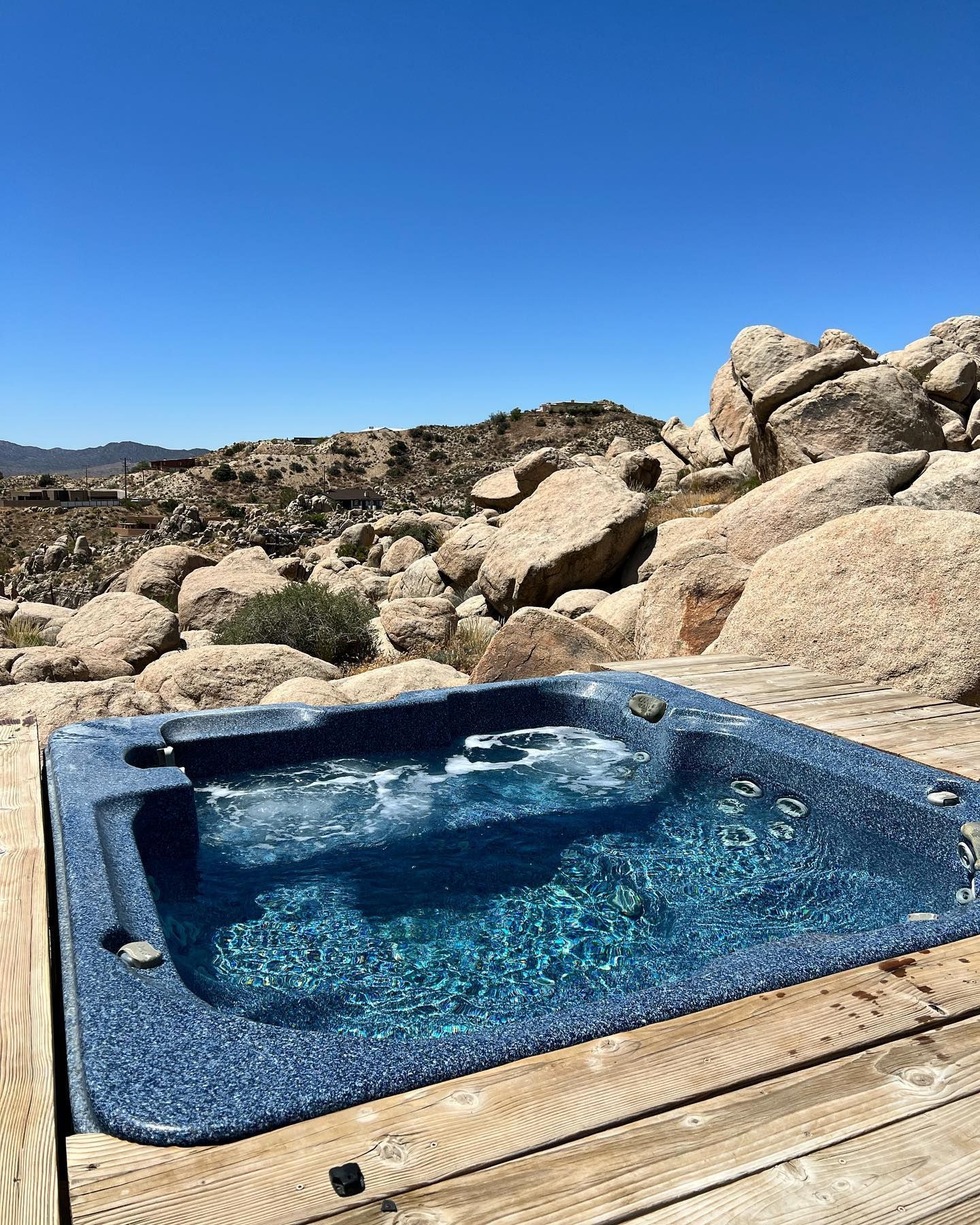 Blue hot tub on wooden deck with a rocky desert landscape in the background, under a blue sky.