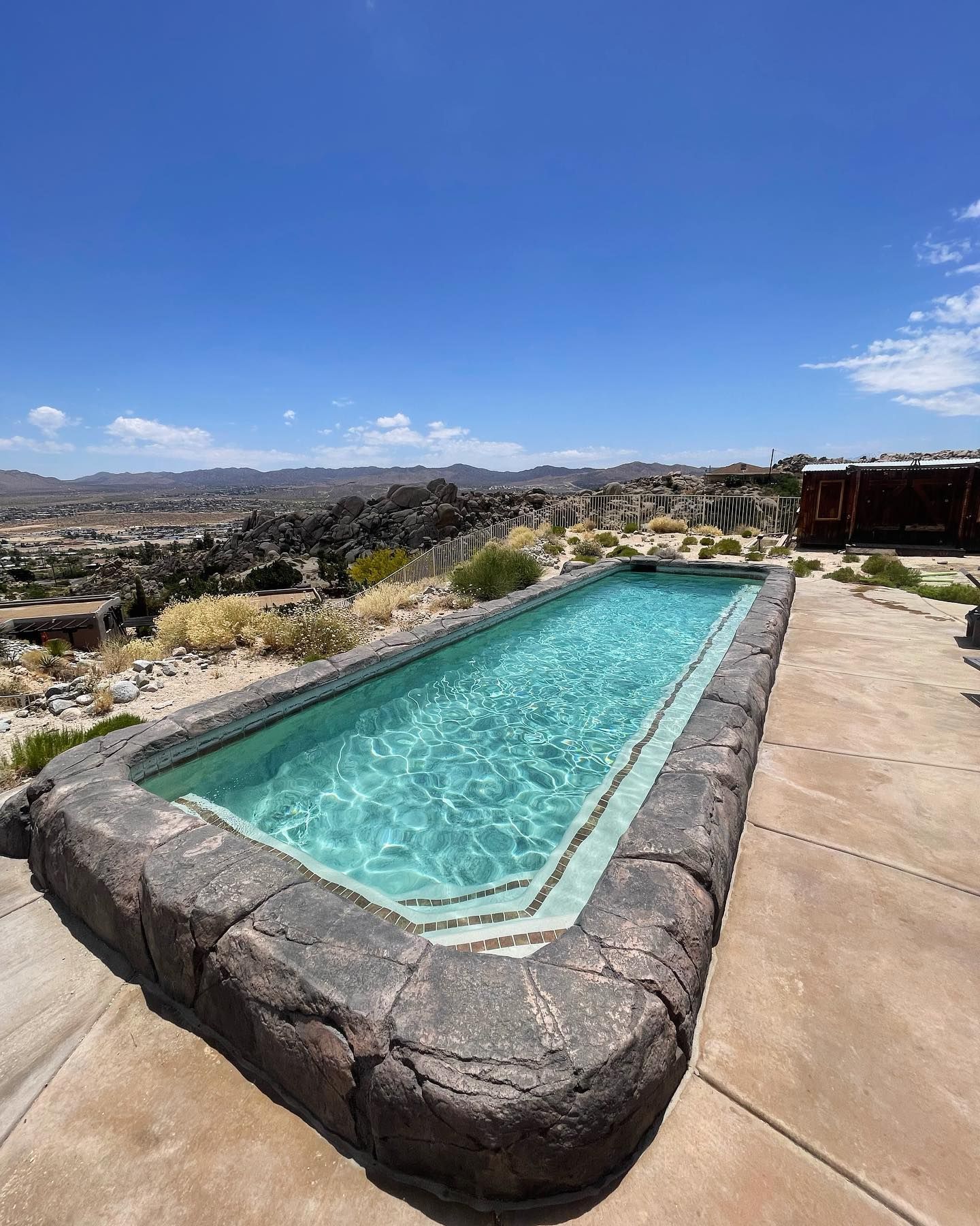 Rectangular swimming pool with rock border, overlooking a desert landscape under a blue sky.