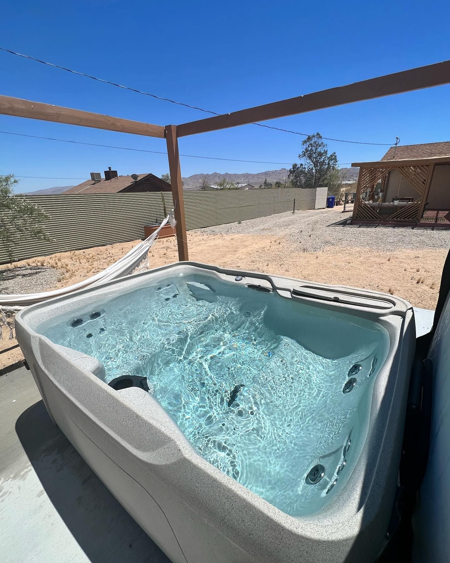 Hot tub filled with water outdoors under a wooden frame, with a metal wall in the background.