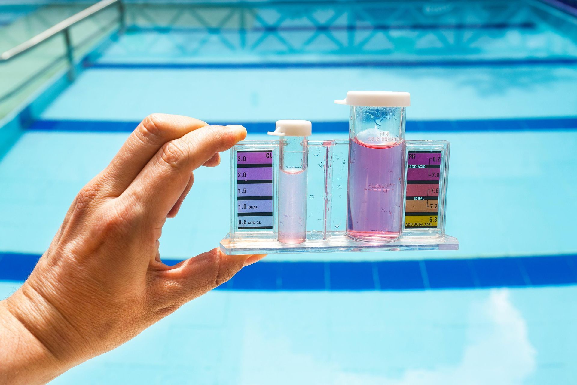 Hand holding a pool water test kit; pink liquid indicates high chlorine in front of a blue pool.