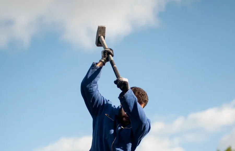 Person in blue coveralls raises a sledgehammer against a cloudy sky.