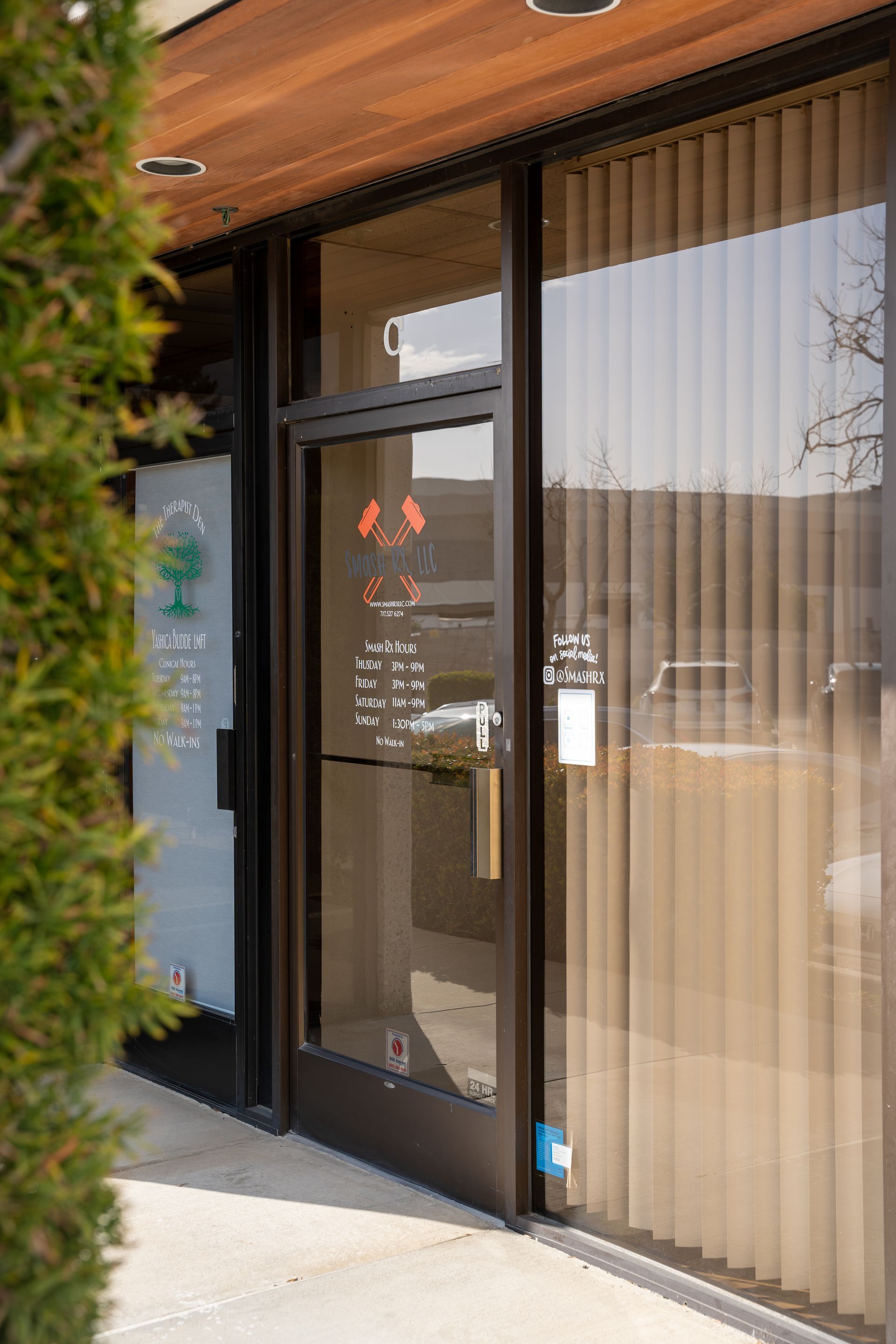Exterior of a business with a glass door, vertical blinds, and a sign. Brown door frame and wooden ceiling.