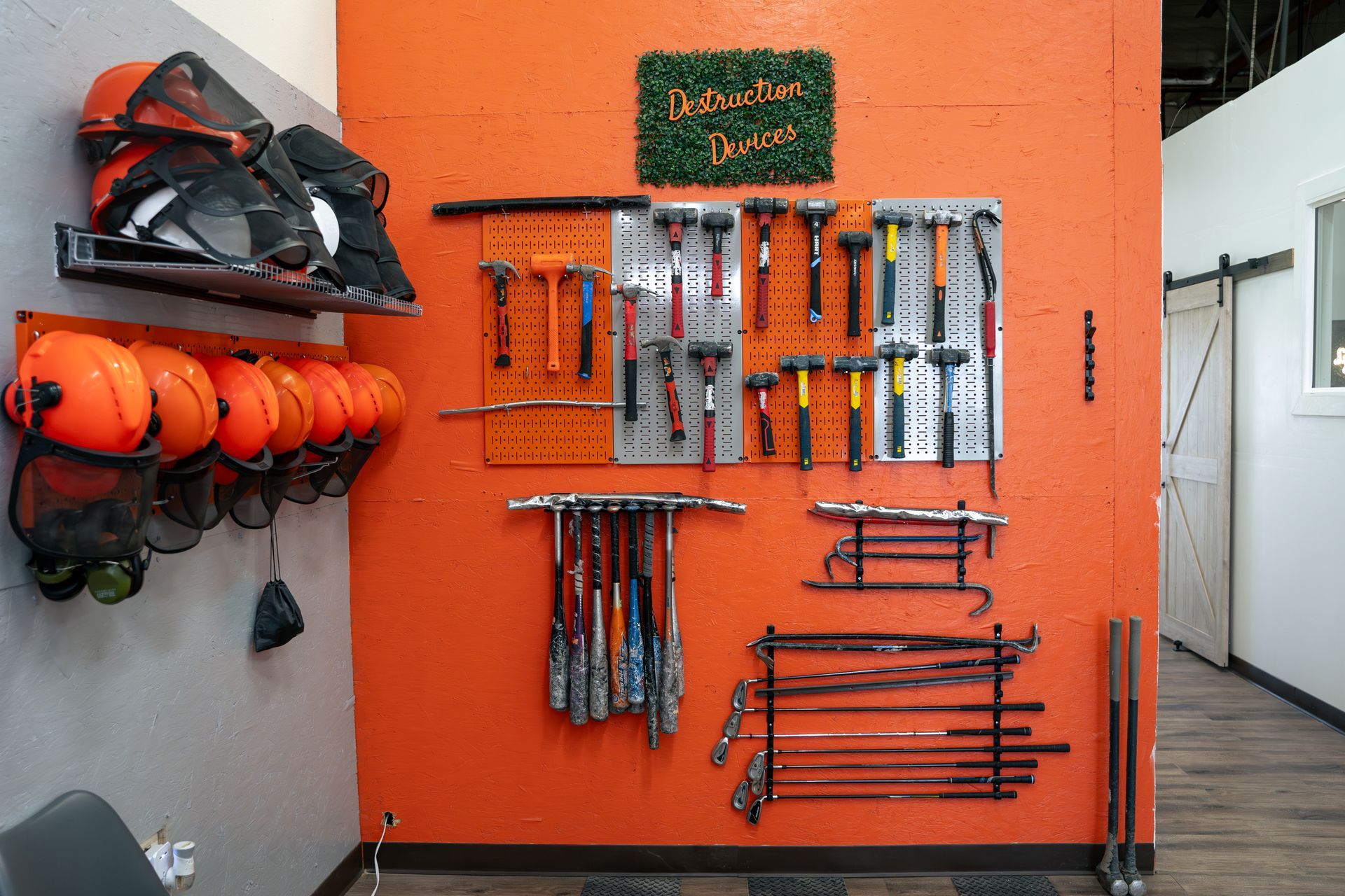 Orange wall displays safety helmets, tools, and bats for axe-throwing or demolition, in a recreational setting.