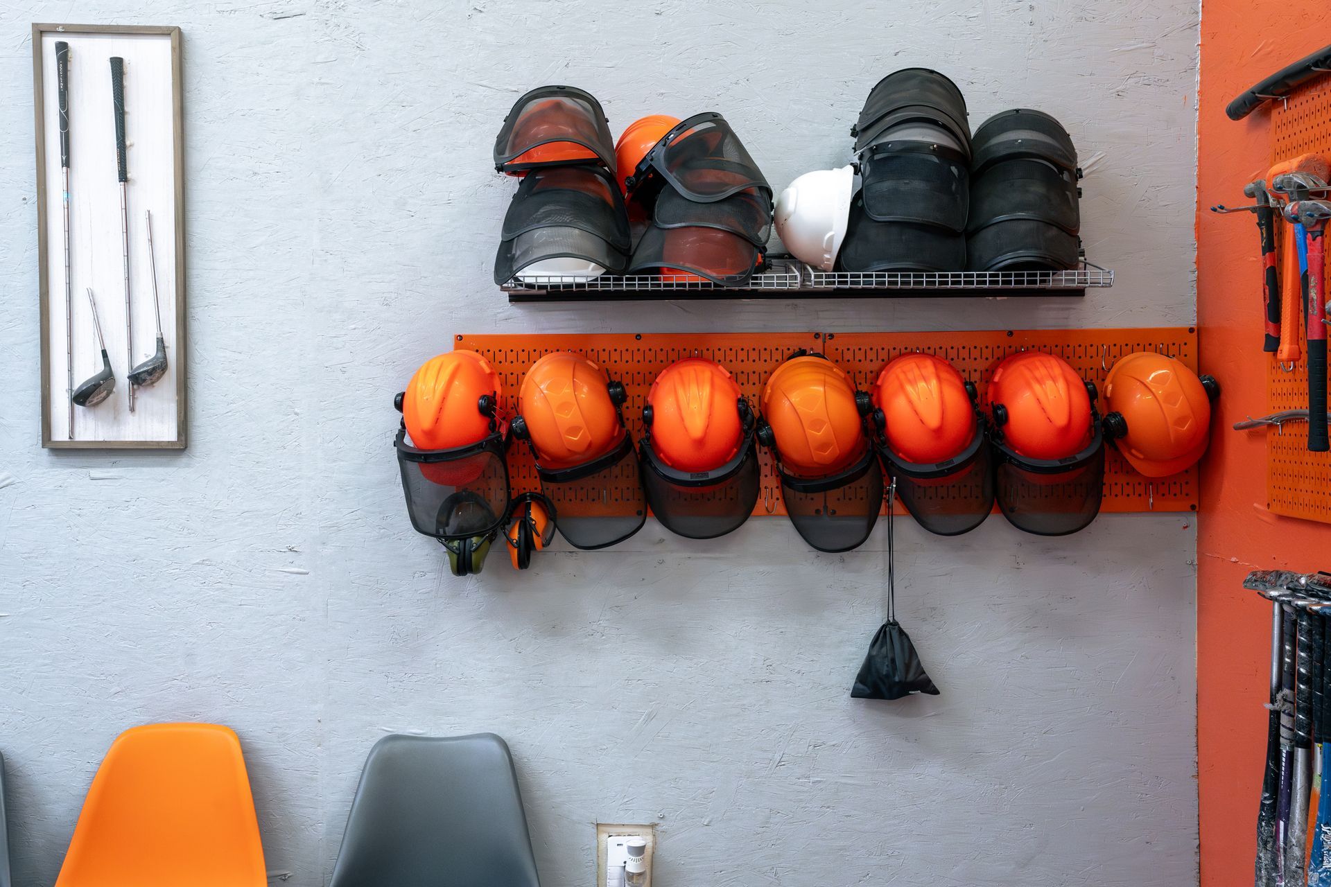 A wall with orange helmets, shoes, and tools; golf clubs on the wall; chairs below.