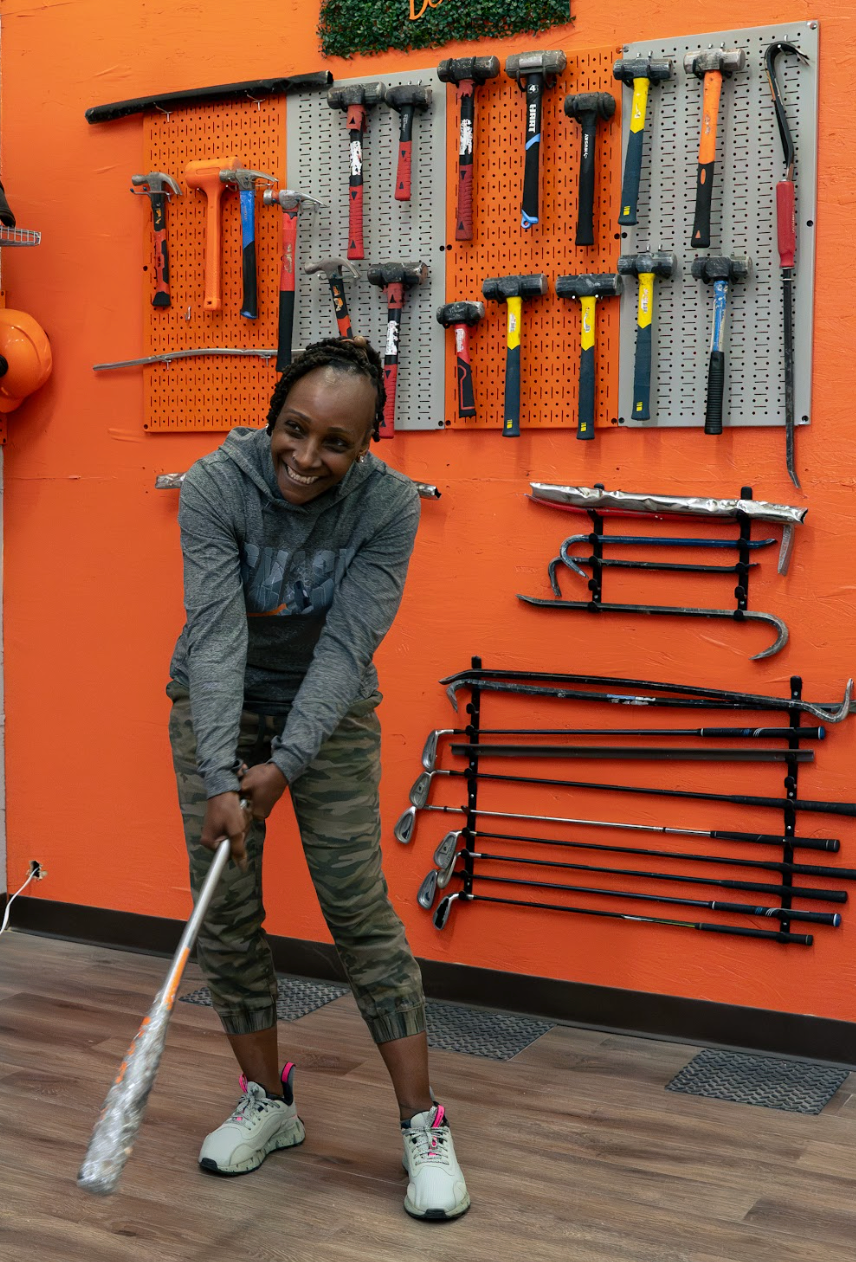 Woman swinging a bat, smiling, in front of a wall of tools. Orange wall background.
