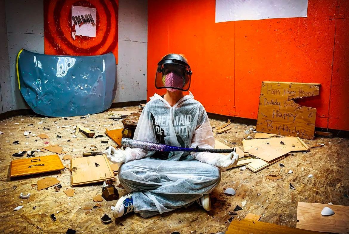 Person in protective gear, holding a bat, sits amid debris in a rage room.