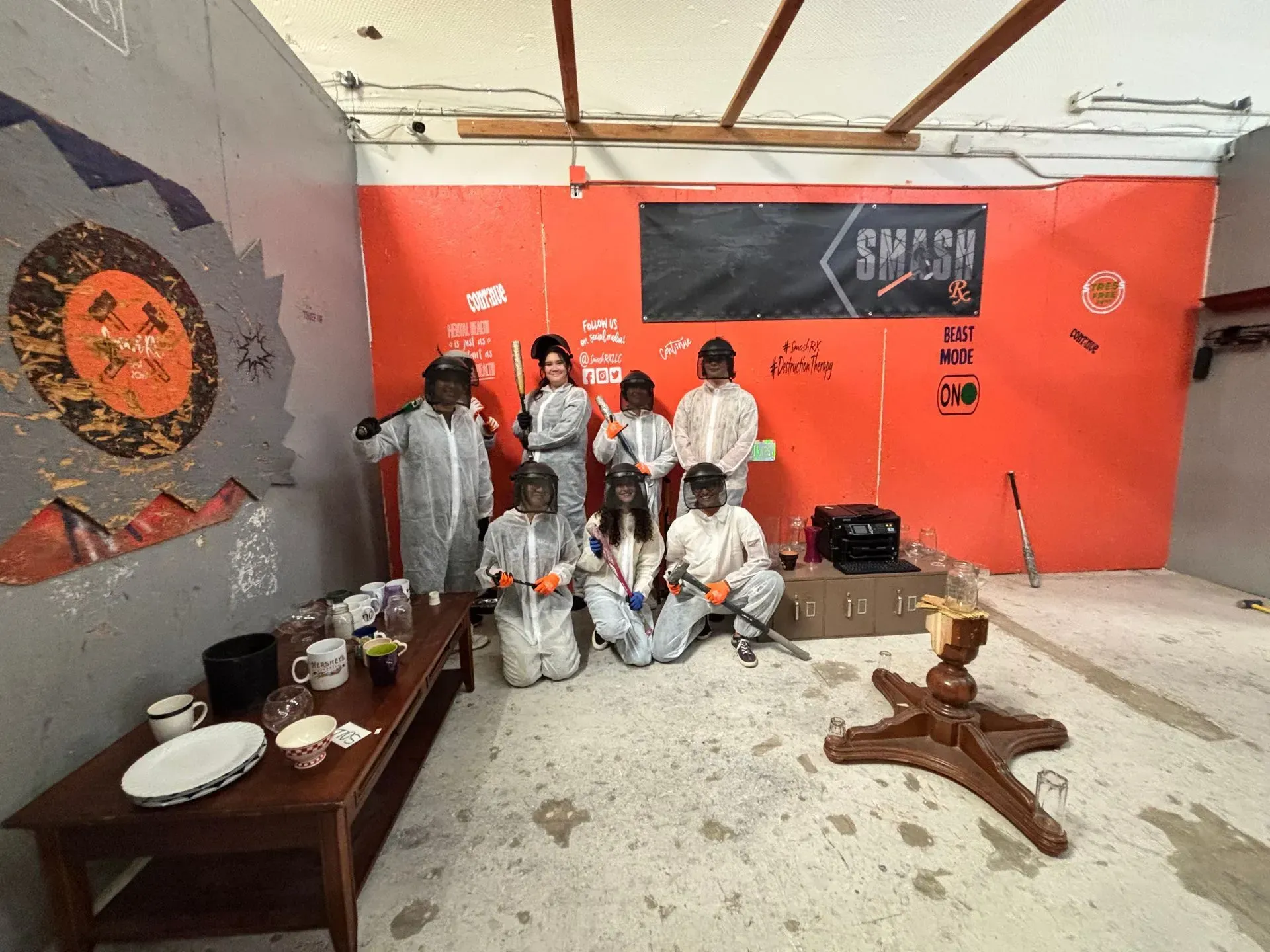 Group of people in protective gear pose in a red-walled smash room.