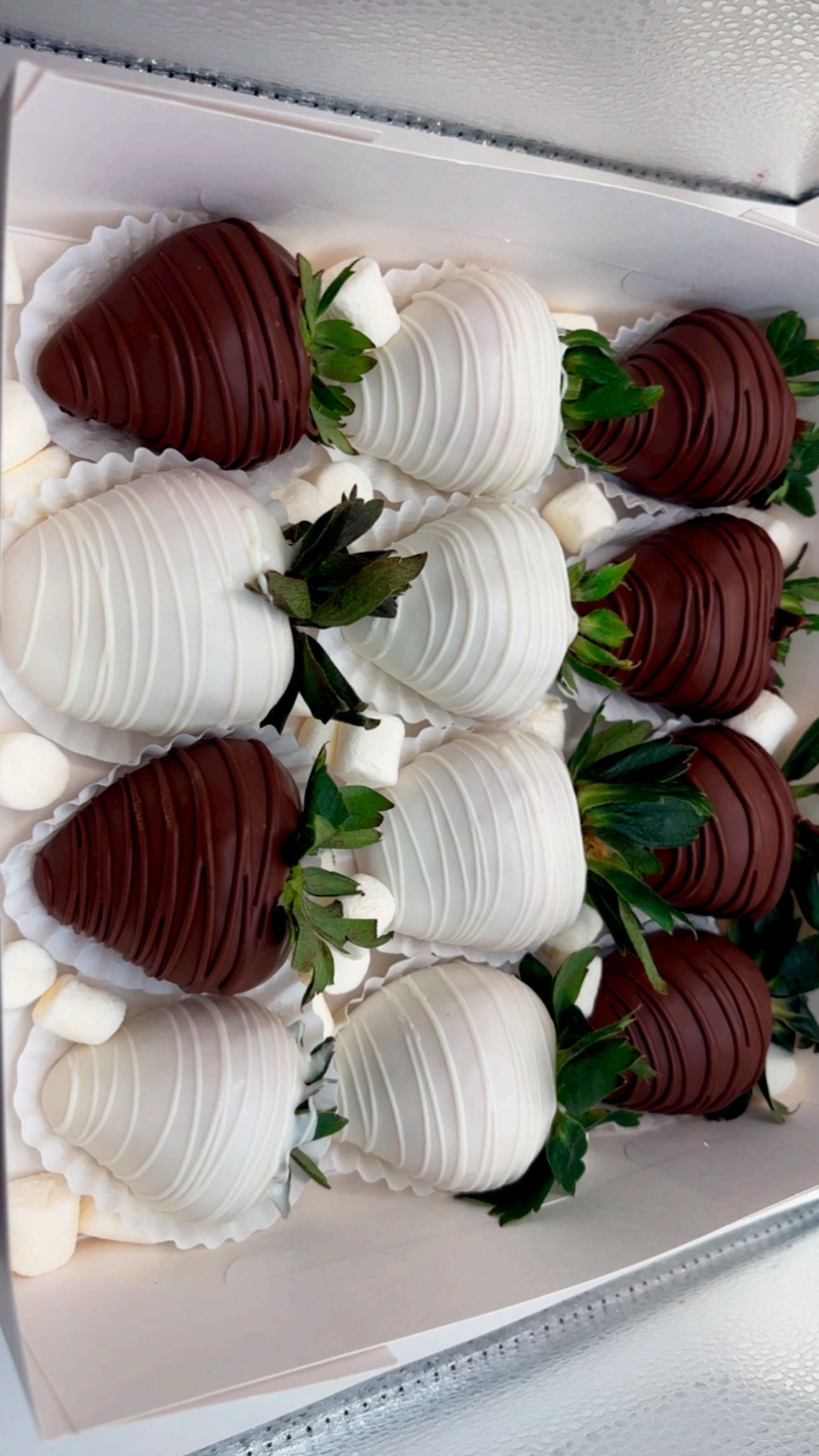 A box of chocolate-covered strawberries, alternating between dark and white chocolate, garnished with small marshmallows.