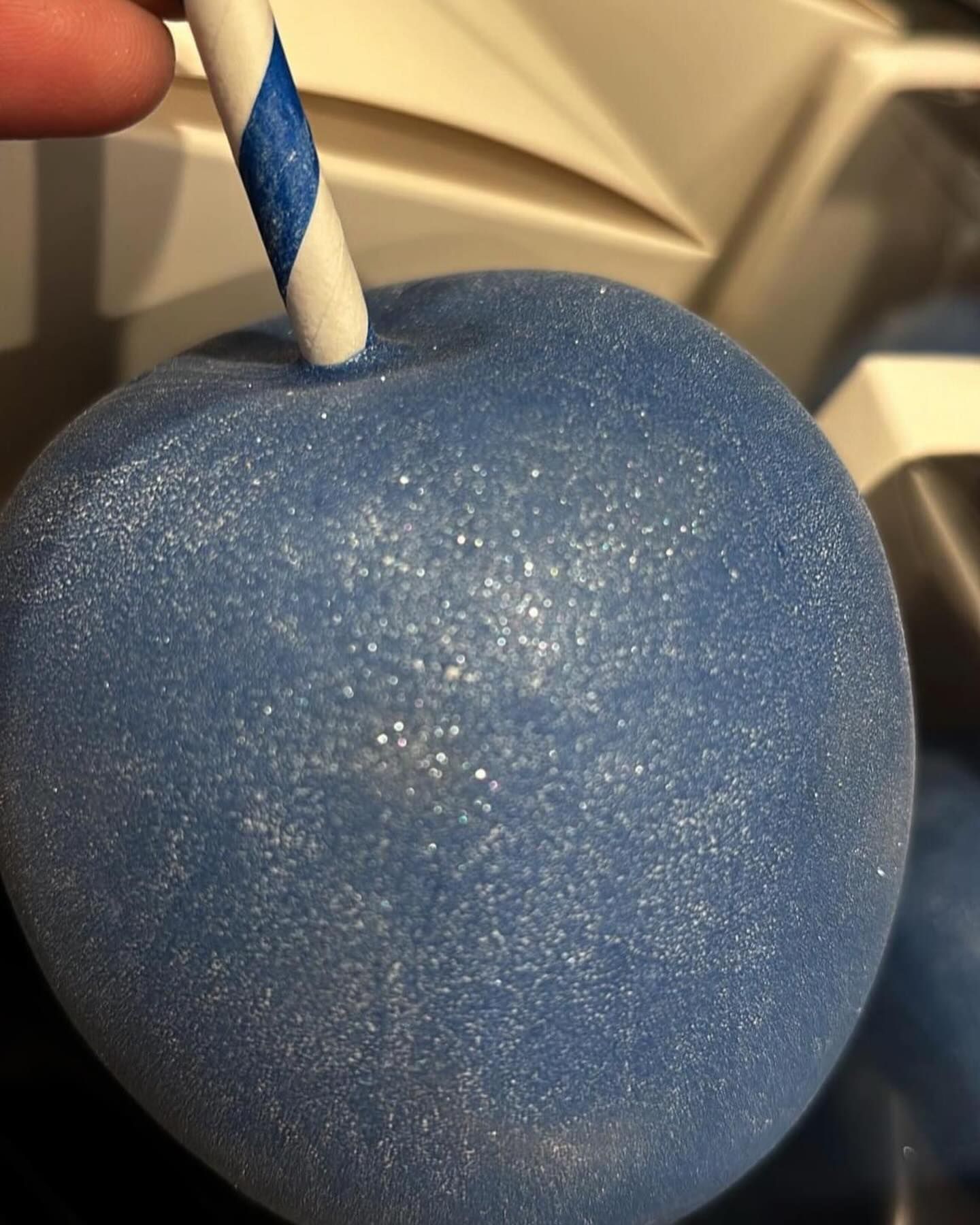 Blue candied apple with white glitter, a blue-striped paper straw inserted into the top.